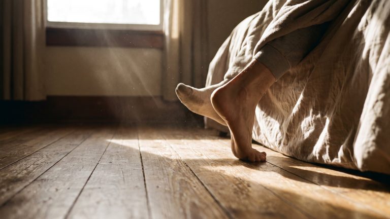 Pie descalzo apoyándose en el suelo de madera junto a la cama a primera hora de la mañana